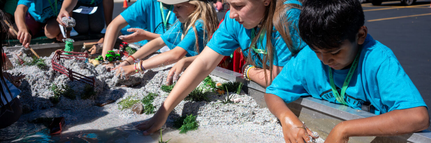 Students in blue shirts gather around a water demonstration table with their hands in the water and sand to learn about watersheds.